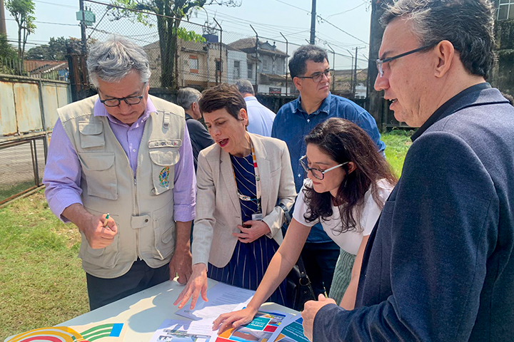 A imagem mostra um grupo de pessoas em uma área externa, aparentemente discutindo um projeto ou plano. Elas estão em volta de uma mesa, analisando papéis e gráficos coloridos