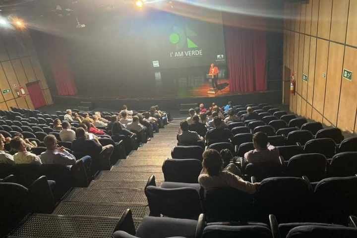 A imagem mostra um auditório ou teatro com fileiras de cadeiras, a maioria delas ocupadas por um público. O palco está à vista, com um palestrante atrás de um pódio no lado direito do palco. Na tela atrás do alto-falante é projetada a logomarca 