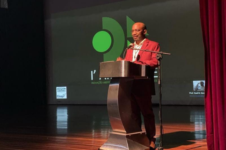 A imagem mostra um homem atrás de um pódio em um palco, fazendo uma apresentação ou discurso. Ele está vestindo um terno vermelho e óculos e parece confiante e engajado com seu público. Atrás dele está um grande telão exibindo o logotipo "I' AM VERDE", incluindo as formas geométricas e a frase "INOVAÇÃO ABERTA PARA MÚLTIPLAS APLICAÇÕES VERDES".  Há também alguns logotipos visíveis na tela, possivelmente representando patrocinadores ou colaboradores do evento, um dos quais parece ser a FINEP, uma organização brasileira focada no financiamento de projetos de ciência, tecnologia e inovação.  Esta configuração indica que o palestrante faz parte de um evento ou conferência formal relacionado à inovação verde, sustentabilidade ou tecnologias ecologicamente corretas, conforme sugerido pelo contexto da iniciativa "I' AM VERDE".