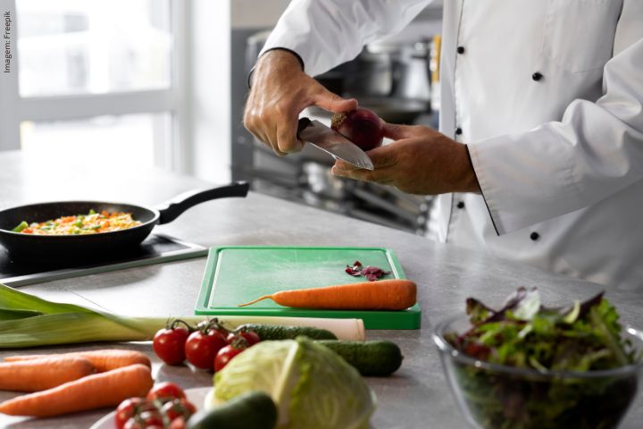 A imagem mostra um chef de cozinha cortando uma cebola roxa em uma tábua de cortar verde. Ao redor da tábua, há vários vegetais frescos, incluindo uma cenoura, tomates cereja, um pepino e folhas de alface. Em segundo plano, uma frigideira no fogão contém o que parece ser uma mistura de vegetais cozidos. A cena parece se passar em uma cozinha profissional.