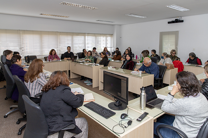 Imagem mostrando uma reunião ou sala de aula. As pessoas estão sentadas em mesas com computadores e notebooks, parecendo envolvidas em discussões ou tomando notas.