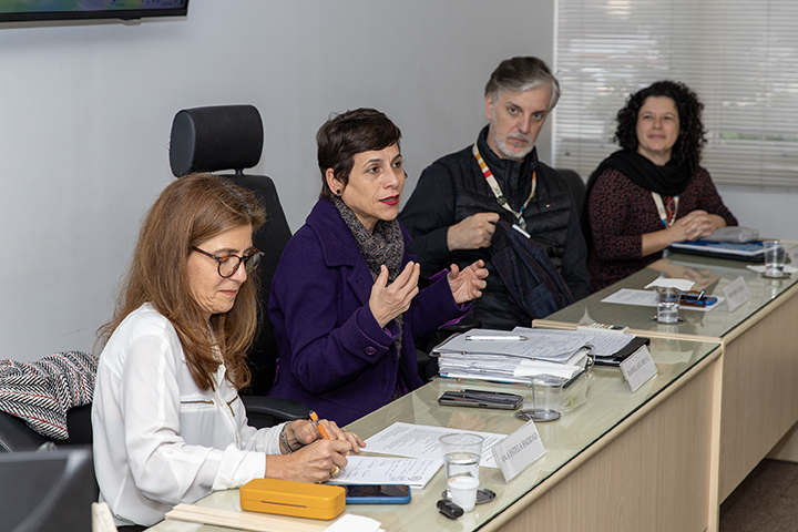 a imagem mostra um grupo de pessoas em um ambiente de reunião formal, com uma mulher falando ao centro (a reitora da Unifesp) enquanto outras ouvem. Placas de identificação e cadernos ficam visíveis na mesa, indicando uma discussão ou apresentação organizada.