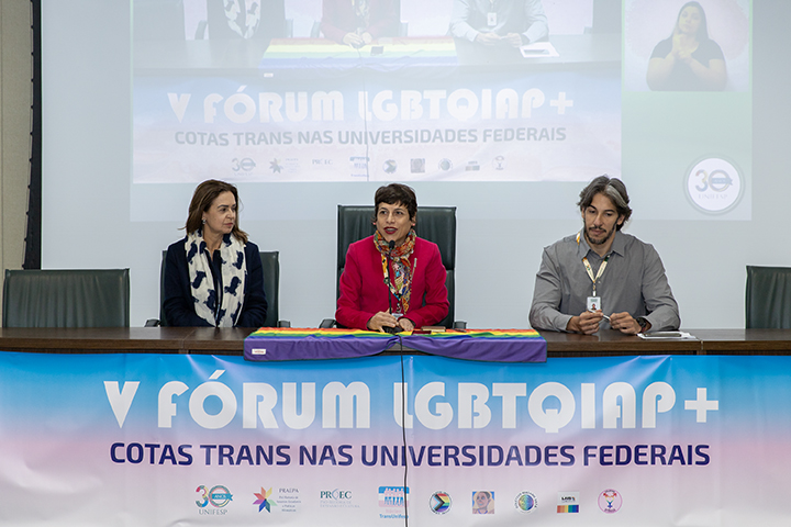 A imagem mostra três pessoas sentadas em uma longa mesa durante um painel de discussão durante o V Fórum LGBTQIAP+, com o banner na frente exibindo o tema do evento: Cotas TRANS nas universidades federais.  A pessoa do meio parece estar falando, vestida com uma jaqueta vermelha, enquanto as outras duas, uma à esquerda e outra à direita, ouvem atentamente. Atrás deles, uma grande tela de projeção exibe novamente o título e o tema do evento, juntamente com os logotipos das organizações envolvidas. O pano de fundo indica que esta é provavelmente uma apresentação formal ou sessão de discussão sobre o tema da inclusão transgênero no ensino superior brasileiro.