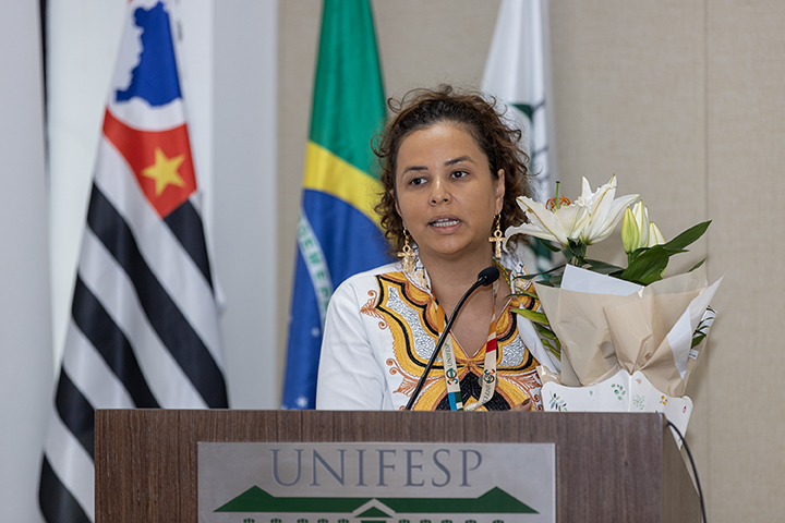 A imagem mostra uma mulher em pé em um pódio rotulado como “UNIFESP”, que significa Universidade Federal de São Paulo. Ela parece estar fazendo um discurso ou apresentação, enquanto fala ao microfone.  Principais detalhes da imagem: - **Pessoa**: A mulher está vestida com uma roupa branca com bordados coloridos, possivelmente de natureza tradicional ou simbólica. Ela tem cabelos cacheados e usa brincos. - **Pódio**: O pódio traz a logomarca da UNIFESP, indicando que o evento provavelmente está ligado à universidade. - **Antecedentes**: Atrás dela há bandeiras, uma delas é a bandeira do Brasil e outra parece ser a bandeira do estado de São Paulo. - **Flores**: Há um buquê de lírios brancos colocado no pódio, sugerindo que o evento pode ser comemorativo, formal ou comemorativo.