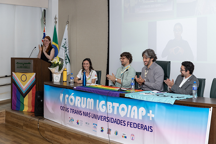 A imagem mostra um painel formal de evento com cinco pessoas sentadas em uma mesa. Um palestrante está à esquerda do painel, batendo palmas enquanto se dirige ao público. O evento parece fazer parte do “V Fórum LGBTQIAP+”, e no banner em frente ao painel está escrito “COTAS TRANS NAS UNIVERSIDADES FEDERAIS”. Isso indica uma discussão focada em aumentar a inclusão de pessoas trans nas universidades federais brasileiras.  Os detalhes na imagem incluem:  Uma grande bandeira de arco-íris e outras bandeiras LGBTQIA+ e inclusivas de gênero estão penduradas sobre a mesa e o pódio. Atrás dos painelistas sentados há duas bandeiras: uma é a bandeira nacional brasileira e a outra parece representar uma instituição. Há uma tela ao fundo mostrando um vídeo ou apresentação, incluindo um intérprete de linguagem de sinais. Os participantes do painel parecem engajados e estão batendo palmas ou sorrindo. A atmosfera da imagem transmite um ambiente positivo e inclusivo, provavelmente focado na promoção dos direitos LGBTQIAP+, especialmente nos direitos trans na educação.