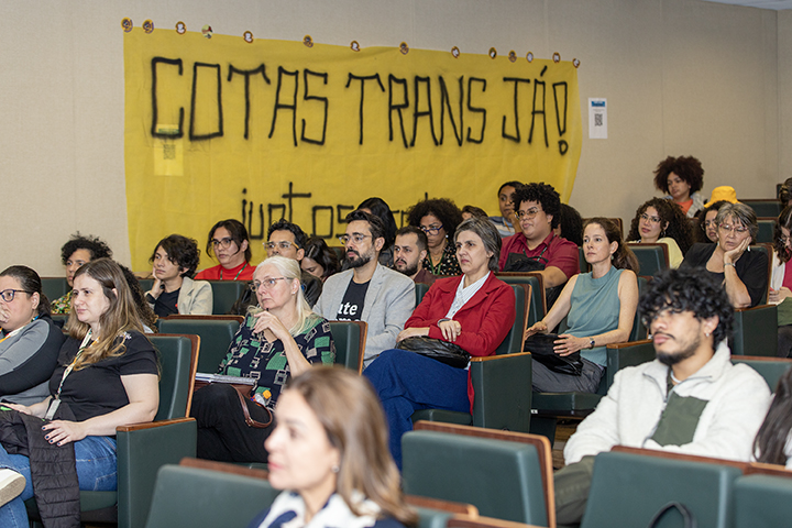 A imagem mostra um público sentado em um auditório ou sala de conferências, participando de um evento no V Fórum LGBTQIAP+. Os participantes são diversos, incluindo pessoas de diversas idades e sexos, todos ouvindo atentamente.  Atrás do público, um grande banner amarelo está pendurado na parede com texto preto em negrito onde se lê Cotas TRANS JÁ! (Cotas trans agora!). Isto indica um forte apoio à implementação imediata de cotas trans nas universidades.