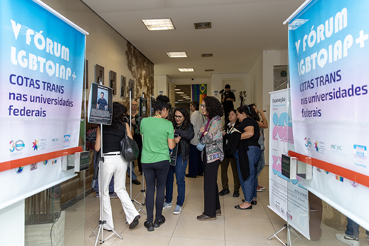 A imagem mostra uma concentração no V Fórum LGBTQIAP+, evento focado nas questões LGBTQIAP+. Dois grandes banners são visíveis em cada lado, com o texto “V Fórum LGBTQIAP+ Cotas TRANS nas universidades federais”, indicando que o evento discute políticas de cotas para transgêneros nas universidades federais.  No centro da imagem, um grupo de pessoas está reunido, aparentemente conversando ou discutindo algo relacionado ao evento. Os participantes parecem diversos e o cenário parece um corredor ou lobby de um prédio, com cartazes e displays espalhados pelas paredes, incluindo fotografias ou painéis informativos. A atmosfera parece casual, mas proposital, com o evento provavelmente sendo educacional ou voltado para ativistas.