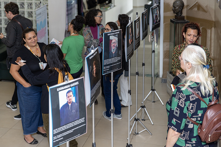 A imagem mostra um grupo de pessoas interagindo no que parece ser uma exposição ou evento. Existem vários pôsteres ou displays montados em estandes, cada um apresentando fotos e descrições de indivíduos, provavelmente apresentando biografias ou perfis. Algumas pessoas estão reunidas em torno dos cartazes, lendo-os ou conversando. O evento parece acontecer em um ambiente fechado, em um espaço bem iluminado e com ambiente casual e amigável.  Várias pessoas estão conversando, algumas olhando para as telas. Uma pessoa com cabelos loiros e uma roupa colorida está focada em um determinado pôster, enquanto outras estão de pé ou conversando nas proximidades. Os próprios pôsteres apresentam retratos e texto, possivelmente destacando profissionais ou indivíduos importantes. 