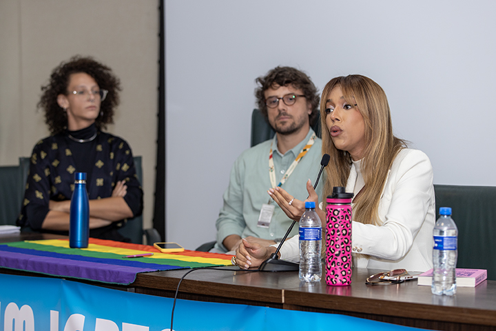 Nesta imagem, três indivíduos estão sentados em um painel, com a pessoa da direita falando ativamente ao microfone. O cenário parece ser uma continuação do evento da imagem anterior, provavelmente parte do “V Fórum LGBTQIAP+”.  Os detalhes incluem:  Palestrante à direita, vestindo um blazer branco, gesticula enquanto se dirige ao público. Na frente deles está uma garrafa de água com estampa rosa e um microfone. Palestrante do meio está de óculos e camisa verde clara, sentado atento e observando o palestrante. Palestrante à esquerda está vestido com um top escuro estampado e tem uma garrafa de água azul na frente dele. Este indivíduo parece atencioso e envolvido com a discussão. A mesa é decorada com uma bandeira do orgulho arco-íris, indicando a temática LGBTQIAP+ do evento. O contexto permanece mínimo, mantendo o foco nos painelistas e na sua discussão. A imagem sugere que o palestrante está discutindo uma questão importante relacionada aos direitos ou políticas LGBTQIAP+, com os demais palestrantes ouvindo atentamente. O cenário continua a refletir um ambiente inclusivo, atencioso e acadêmico.