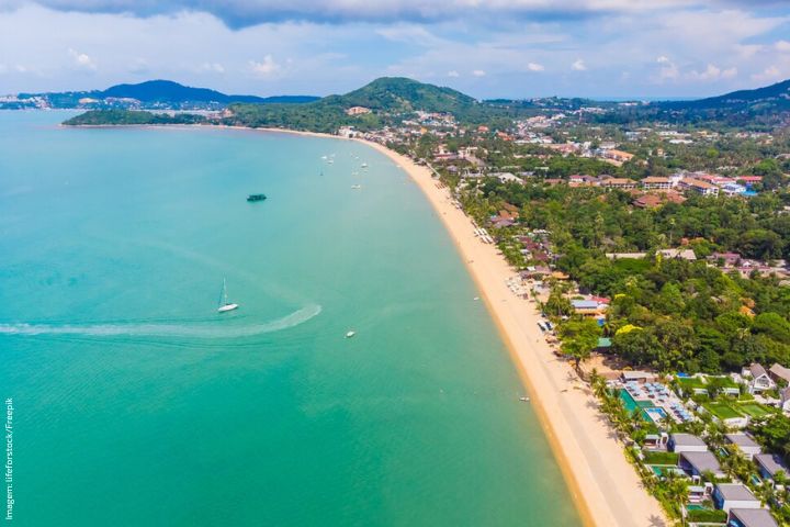 A imagem mostra uma vista aérea de uma linha costeira. A praia é longa e de areia clara, margeada por um mar de águas azul-turquesa. À direita, há uma faixa de vegetação verde densa, intercalada com construções, que se estende ao longo da costa. A região parece urbanizada, com várias casas e possivelmente alguns edifícios. No mar, perto da costa, há algumas embarcações. Ao fundo, colinas verdes se erguem, complementando a paisagem costeira. O céu está parcialmente nublado, indicando um dia claro com algumas nuvens.