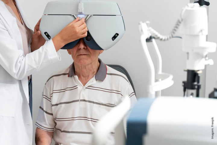 A imagem mostra um homem sendo submetido a um exame oftalmológico com equipamento oftalmológico avançado. O homem, provavelmente um paciente, está sentado enquanto um profissional de saúde ajusta um dispositivo grande e moderno diante de seus olhos. A máquina, que cobre ambos os olhos, parece ser um dispositivo de teste óptico, comumente usado em exames oftalmológicos para medir a visão ou diagnosticar doenças oculares. O ambiente é um consultório clínico ou médico, com equipamento de diagnóstico adicional visível ao fundo. A imagem sugere um exame oftalmológico de rotina ou uma avaliação especializada da visão.