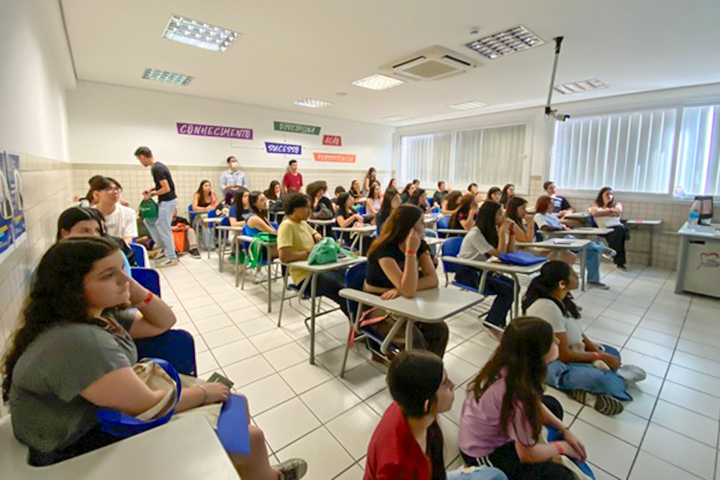 A imagem mostra um ambiente semelhante a uma sala de aula, onde muitos alunos ou participantes estão sentados, alguns em carteiras e outros no chão. A sala é bem iluminada com paredes brancas e grandes janelas que permitem a entrada de luz natural. Há placas nas paredes com palavras como “CONHECIMENTO”, “DISCIPLINA”, “FOCO”, “SUCESSO” e “AÇÃO”, que são termos motivacionais em português, que significam “conhecimento”, “disciplina”, “foco”. "sucesso" e "ação". Parece ser uma sala de aula ou um evento que envolve aprendizagem ou uma apresentação, possivelmente uma exposição ou evento educacional.