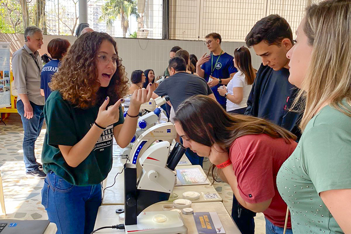 Nesta imagem, há uma animada exposição ou workshop científico acontecendo, provavelmente em um ambiente educacional. Várias pessoas estão reunidas em torno de mesas com microscópios e outros equipamentos científicos. No lado esquerdo, uma jovem de cabelos cacheados, vestindo uma camiseta verde e óculos, parece estar explicando algo entusiasticamente para um grupo. Sua expressão facial e gestos com as mãos sugerem que ela está envolvida em uma conversa ou demonstração. No meio da imagem, outra mulher vestindo uma camisa vermelha está se inclinando para olhar através de um microscópio. Há algumas outras pessoas ao redor das mesas, possivelmente estudantes, prestando atenção às exposições e demonstrações. O fundo mostra mais pessoas envolvidas em atividades semelhantes, indicando que pode ser uma feira de ciências, exposição ou evento educacional. O local parece ser interno, possivelmente em uma escola ou universidade, pois há várias pessoas de diferentes idades presentes.