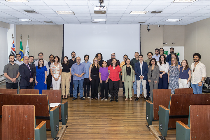 A imagem mostra um grupo de pessoas reunidas em um ambiente formal, possivelmente uma sala de conferências ou auditório, com cadeiras de madeira e uma grande tela branca ao fundo.  No centro da imagem, estão alinhadas aproximadamente 24 pessoas, sugerindo que se trata de um evento de caráter oficial ou institucional. A maioria está vestida de maneira casual ou semi-formal, e a atmosfera parece ser de celebração ou registro de um momento importante.  À esquerda, pode-se observar bandeiras, incluindo a bandeira do estado de São Paulo e do Brasil, reforçando que o evento provavelmente ocorreu em uma instituição pública brasileira. Considerando o contexto e as vestimentas, o grupo poderia ser composto por docentes, servidores ou pesquisadores participando de uma cerimônia de posse ou de reconhecimento.