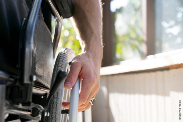 A imagem mostra um close da mão de uma pessoa segurando o volante de uma cadeira de rodas. A pessoa parece estar impulsionando manualmente a cadeira de rodas, com parte do braço visível. A luz natural sugere que o ambiente pode ser interno, perto de uma janela. A imagem transmite mobilidade e independência, com foco no ato de movimento.