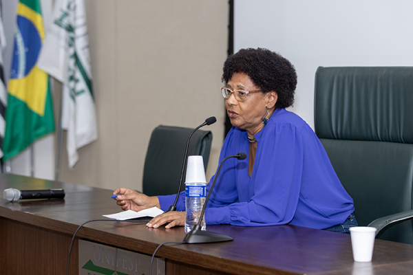 A imagem mostra uma mulher negra sentada a uma mesa, falando ao microfone no que parece ser um evento formal, possivelmente uma palestra ou conferência. Ela está vestindo uma blusa azul e óculos, e tem cabelos curtos e cacheados. Na frente dela estão papéis, uma garrafa de água e um copo pequeno.  Atrás dela há um muro com diversas bandeiras, inclusive uma que parece ser do Brasil, baseada nas cores visíveis verde, amarelo e azul. O cenário lembra um espaço acadêmico ou institucional, tendo como pano de fundo parte da logomarca da “UNIFESP”, sigla para Universidade Federal de São Paulo no Brasil.  O palestrante está em uma postura focada e profissional, provavelmente fazendo uma palestra ou apresentação.