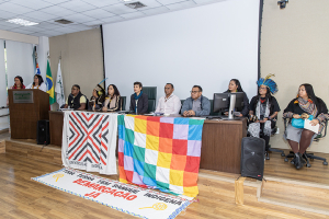 Esta imagem mostra um painel de pessoas sentadas no palco de um evento, provavelmente um seminário ou conferência. O painel inclui homens e mulheres, e alguns deles usam trajes tradicionais indígenas, como cocares de penas, indicando um enfoque cultural ou indígena.  Dois banners coloridos estão pendurados na frente dos palestrantes. Um banner diz &quot;Universidade Indígena&quot; e o outro é um padrão xadrez multicolorido, frequentemente associado a movimentos indígenas. Há também um grande pano no chão com a frase “Tem Sangue Indígena – Demarcação Já”, que se traduz como “Há Sangue Indígena – Demarcação Agora”, provavelmente um apelo aos direitos e reconhecimento das terras indígenas.  A sala é um ambiente formal com bandeiras brasileiras e bandeiras institucionais visíveis ao fundo, sugerindo que está acontecendo em um local acadêmico ou governamental. O evento parece estar centrado em questões, direitos, educação ou preservação cultural indígenas. As pessoas no pódio e no palco possivelmente estão discutindo esses temas.